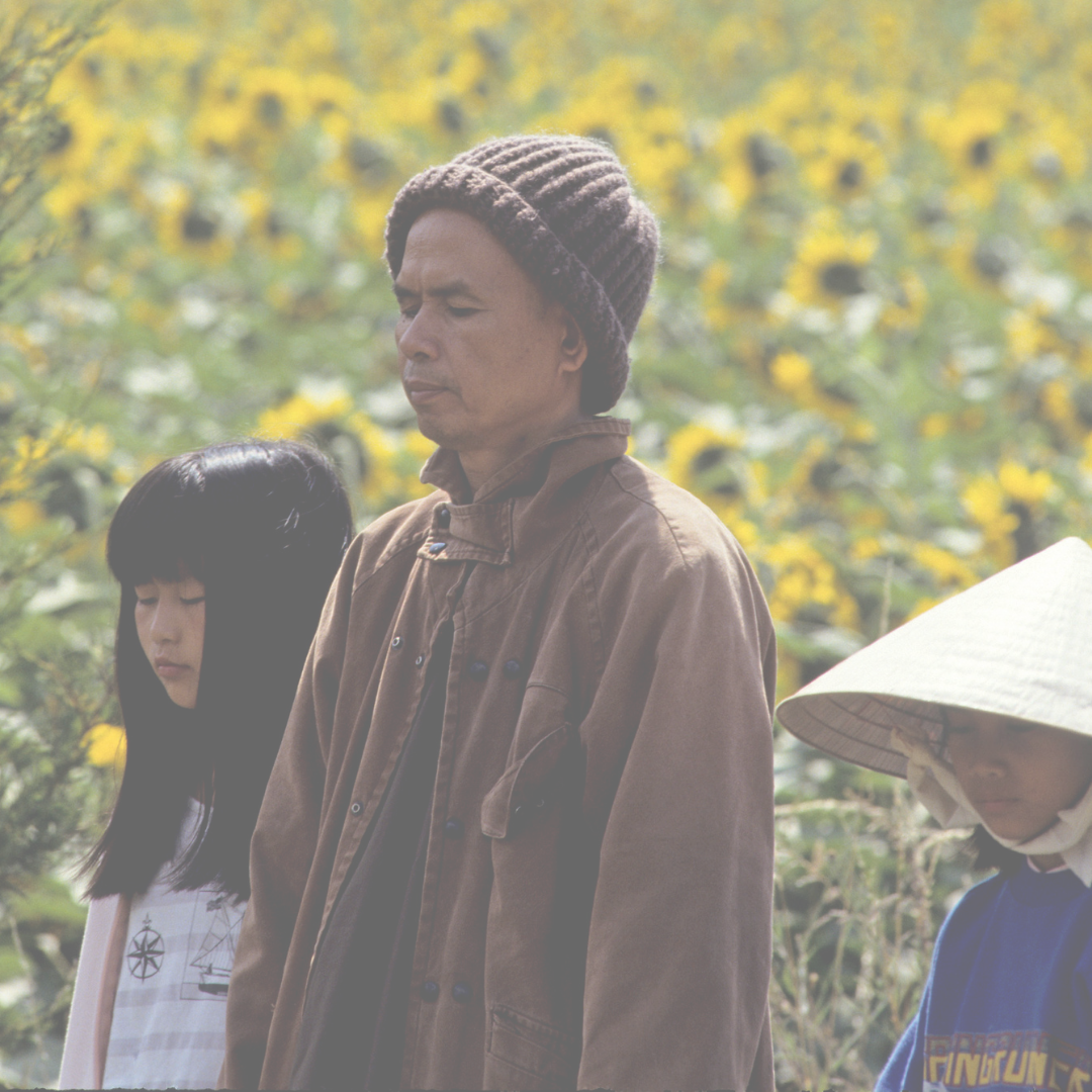 Thich Nhat Hanh and two children standing in a field of sunflowers.